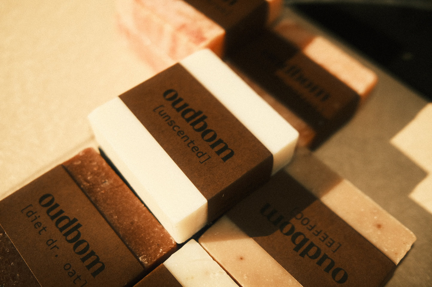 Photo of a white bar of soap with a brown “oudbom, unscented” label, atop various other bars of labeled soaps 