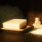 White bar of soap on light brown soap dish, a pile of sand to its side with a stack of small cubes behind. Brown background and floor