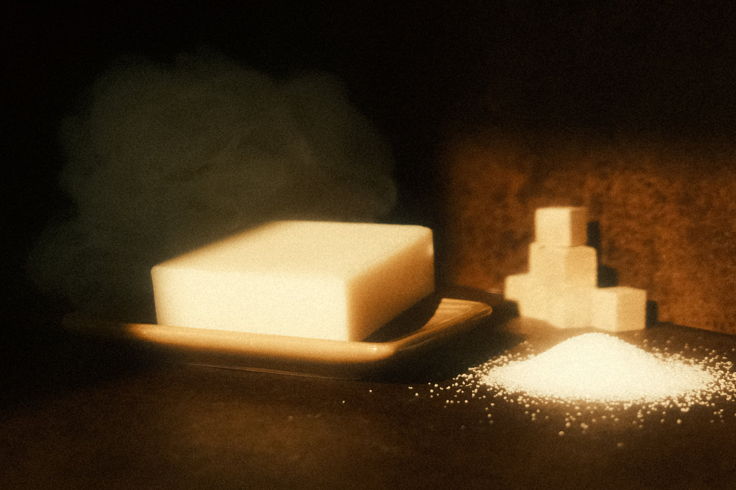 White bar of soap on light brown soap dish, a pile of sand to its side with a stack of small cubes behind. Brown background and floor