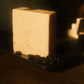 A tan bar of soap standing on a black soap dish with a small pile of coffee beans, brown background