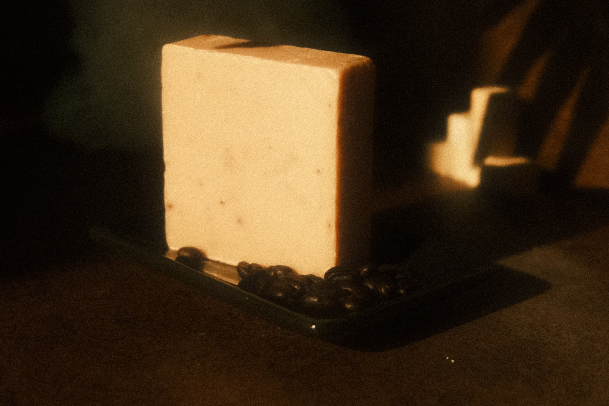 A tan bar of soap standing on a black soap dish with a small pile of coffee beans, brown background