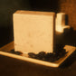 A tan bar of soap stands on a light brown soap dish with coffee beans surrounding it