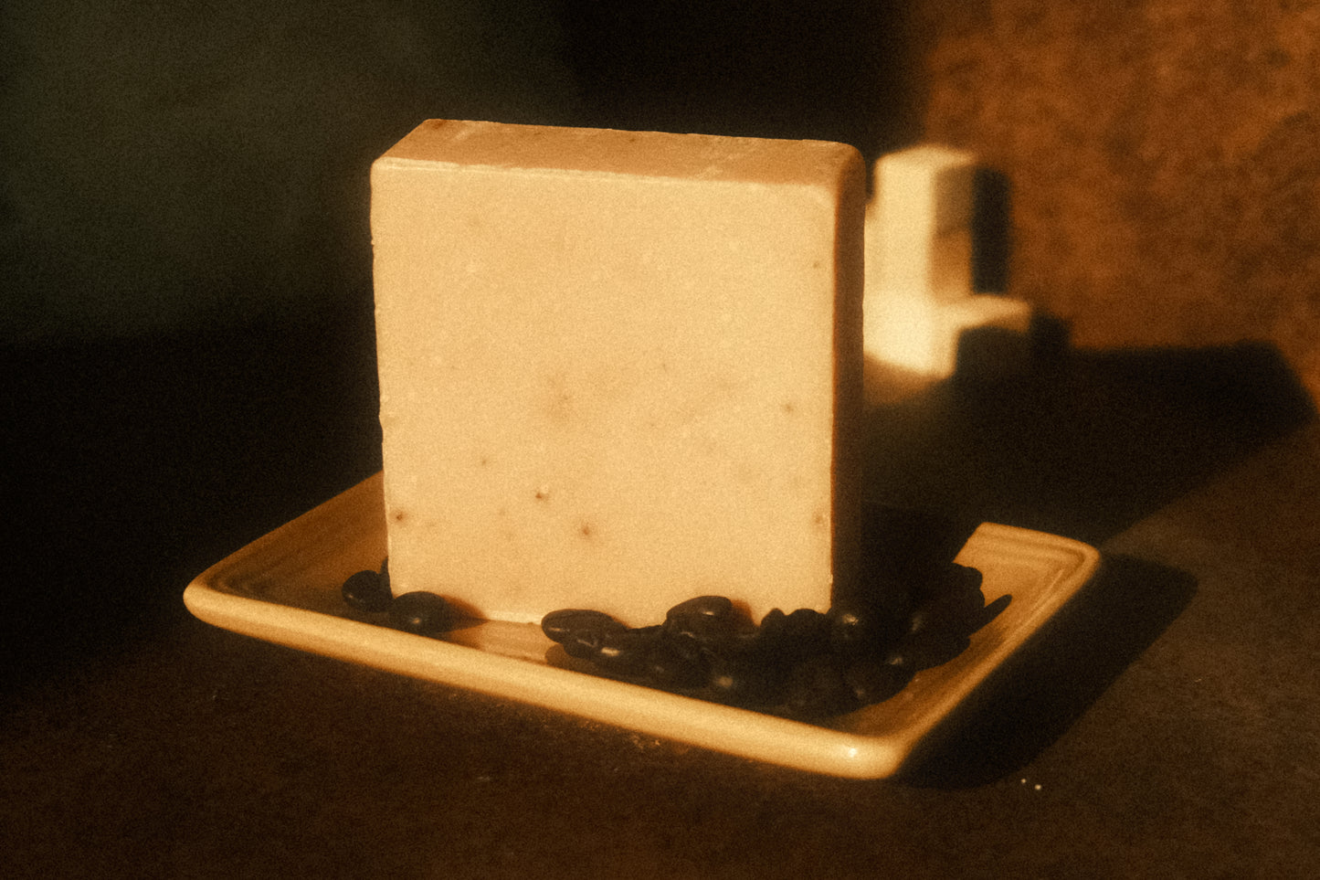 A tan bar of soap stands on a light brown soap dish with coffee beans surrounding it