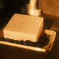 Tan bar of soap laying on a soap dish with coffee beans surrounding it, a stack of cubes in the background