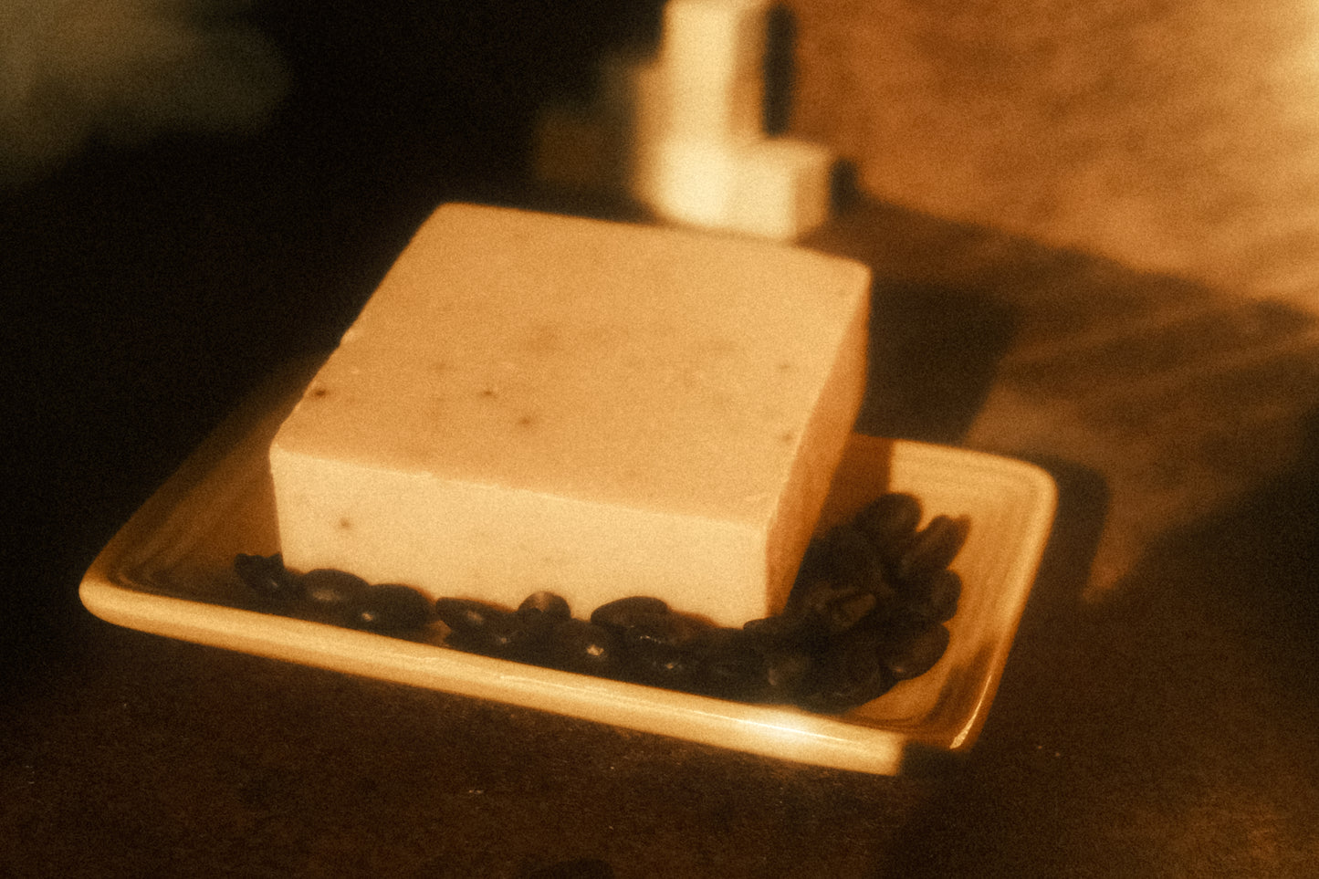 Tan bar of soap laying on a soap dish with coffee beans surrounding it, a stack of cubes in the background