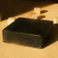 Black bar of soap laying flat on beige floor with small cubes behind it