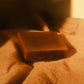 Brown bar of soap laying flat on beige cloth, a third of the photo within a shadow