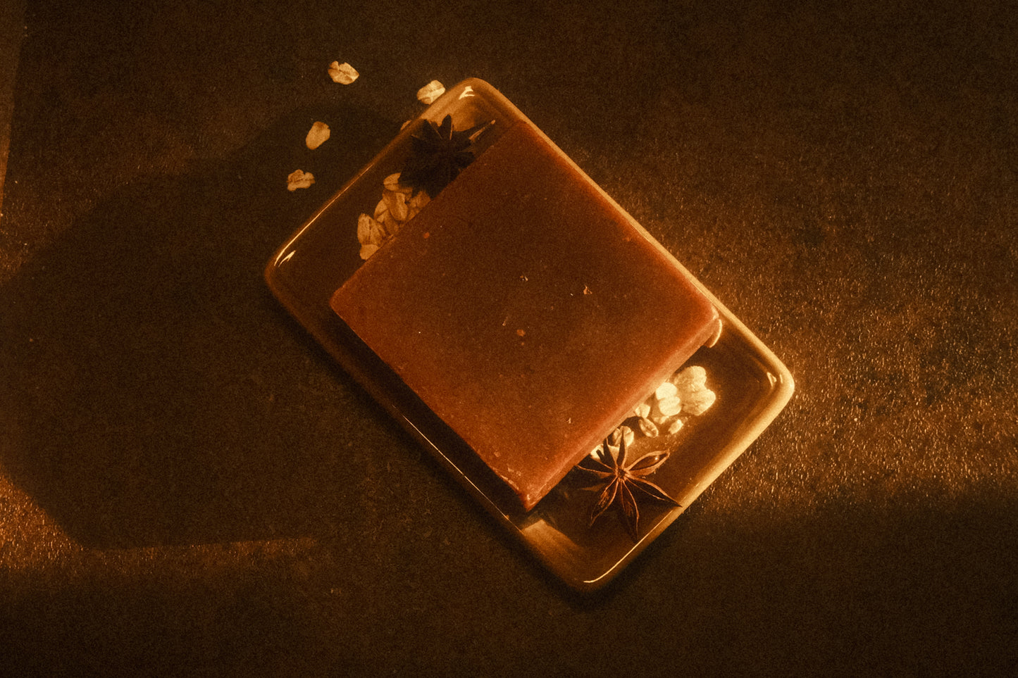 Top down view of a brown bar of soap laying on a tan soap dish with oats and star anise garnishing it