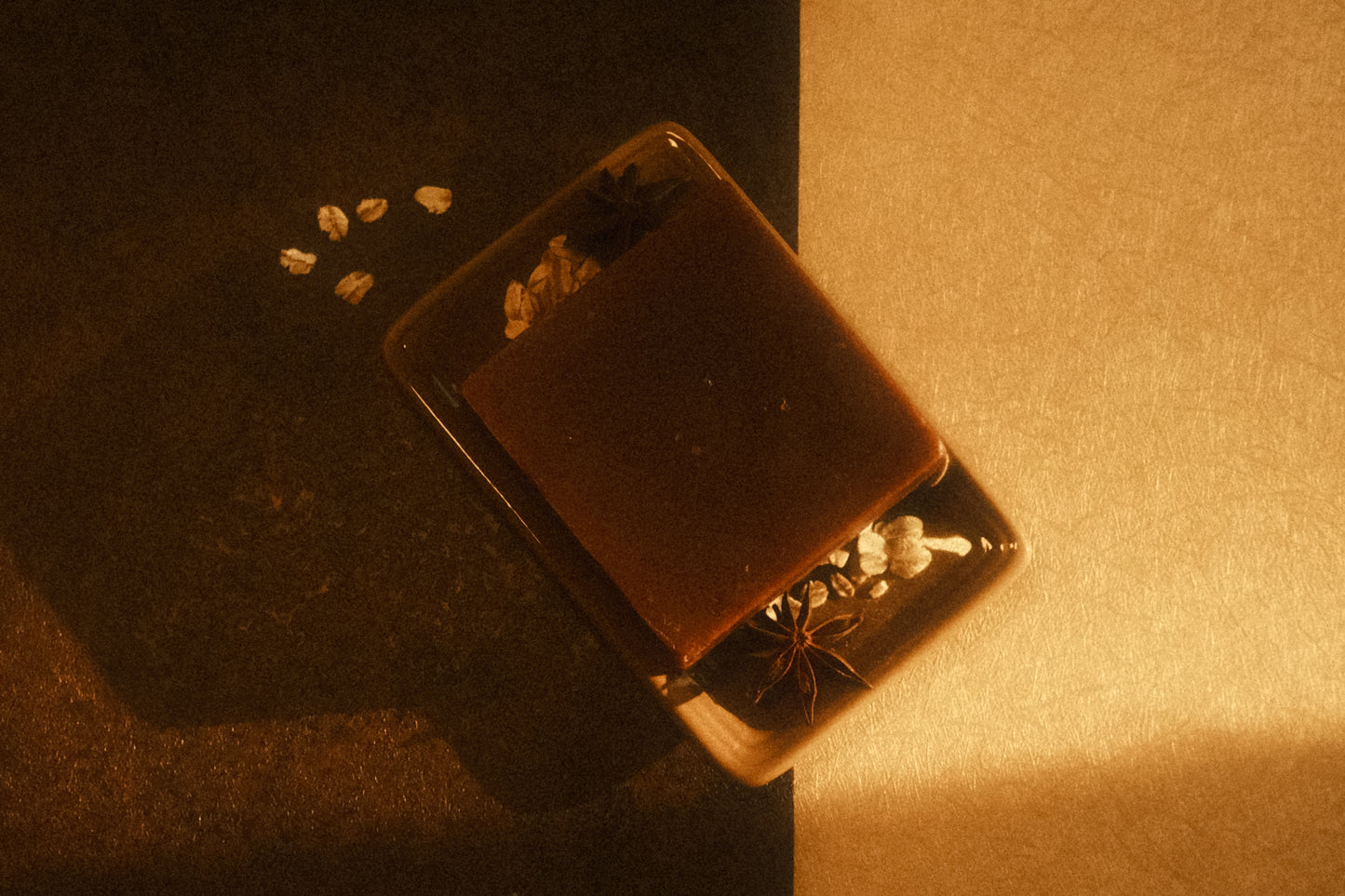 Top down view of a brown bar of soap laying on a tan soap dish with oats and star anise garnishing it