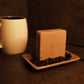 Tan bar of soap standing on light brown soap dish with coffee beans surrounding it. A coffee mug sits int he background