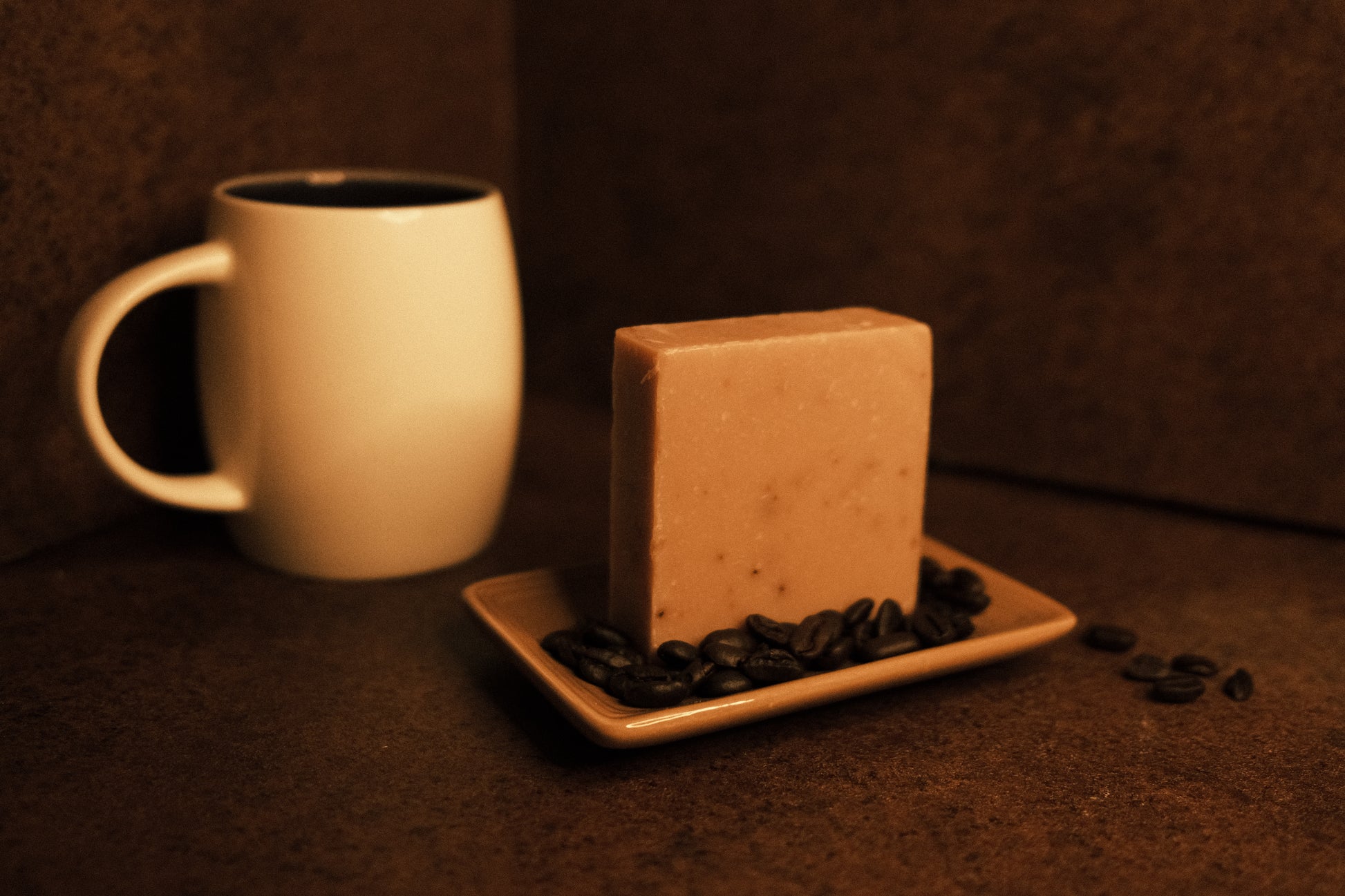 Tan bar of soap standing on light brown soap dish with coffee beans surrounding it. A coffee mug sits int he background