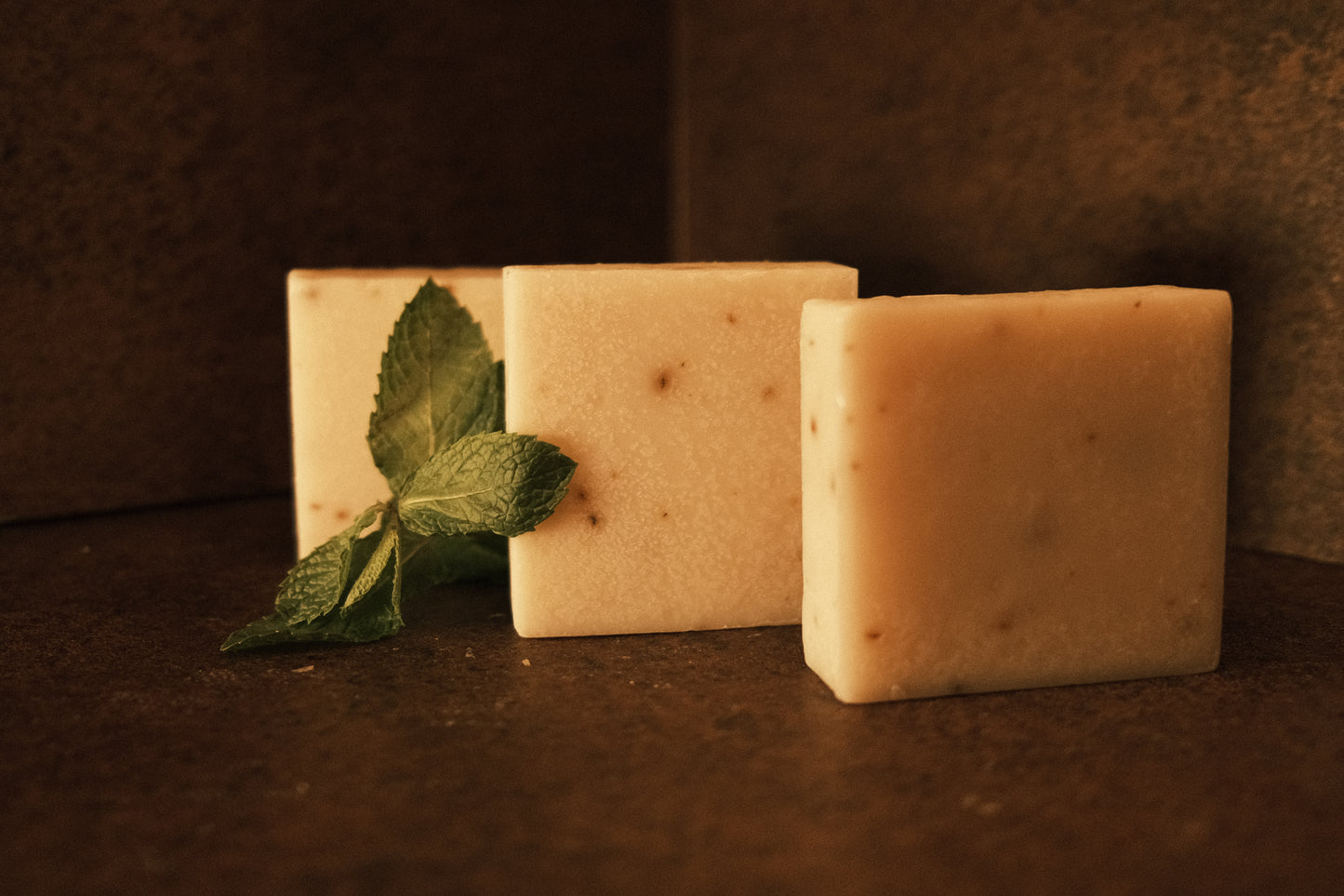 Three bars of white soap stand in ascending order with mint leaves garnishing the side, brown background and floor