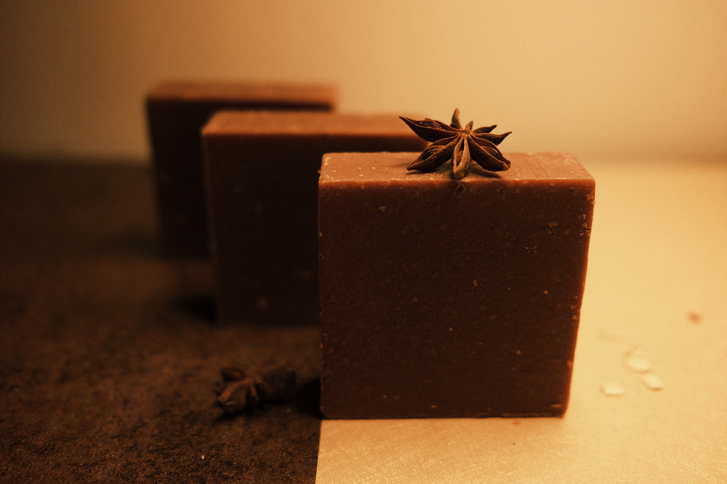 Three brown bars of soap standing in ascending order with half brown and half beige floor, star anise on top of the front bar and to its side.