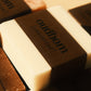 Close up shot of a white bar of soap with a brown “oudbom, unscented” label, atop various other bars of labeled soaps 