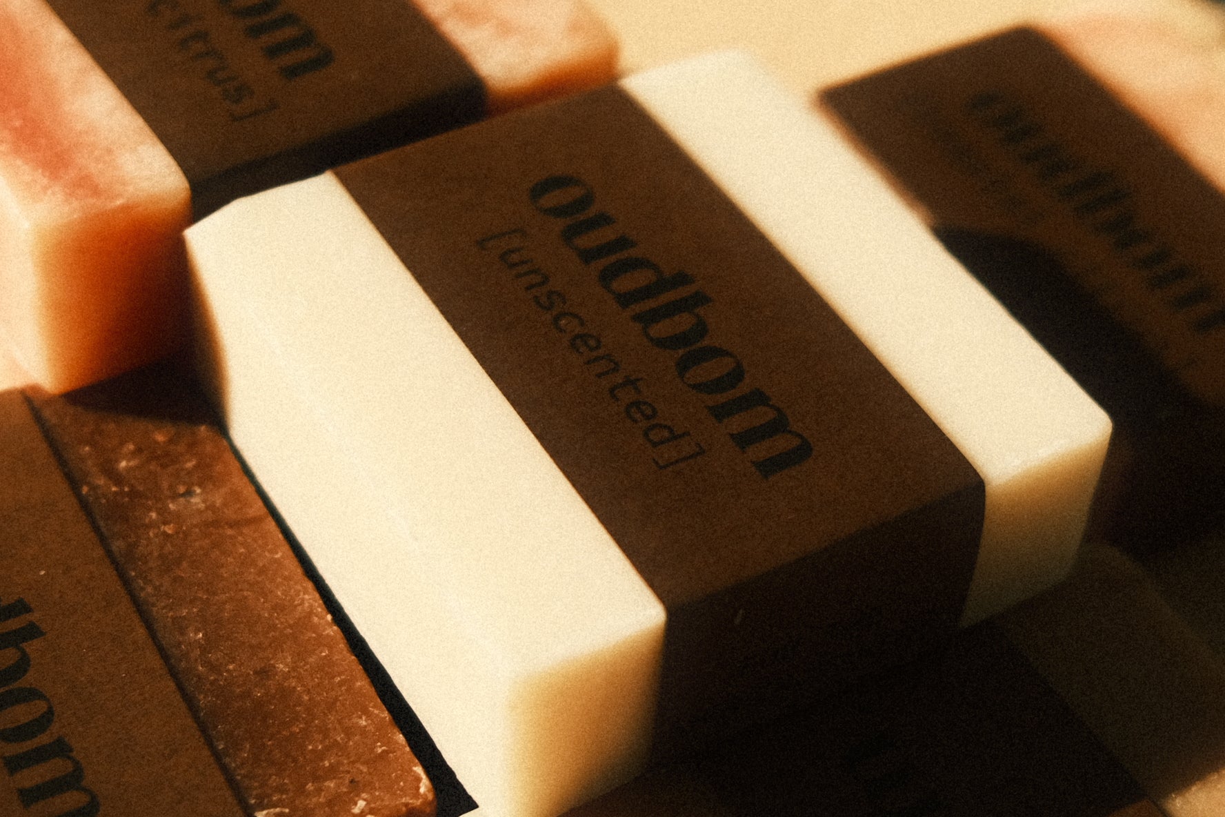 Close up shot of a white bar of soap with a brown “oudbom, unscented” label, atop various other bars of labeled soaps 